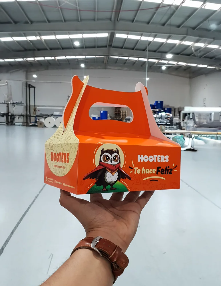 La caja de menú infantil de Hooters sostenida en la mano, en la planta de Compospack